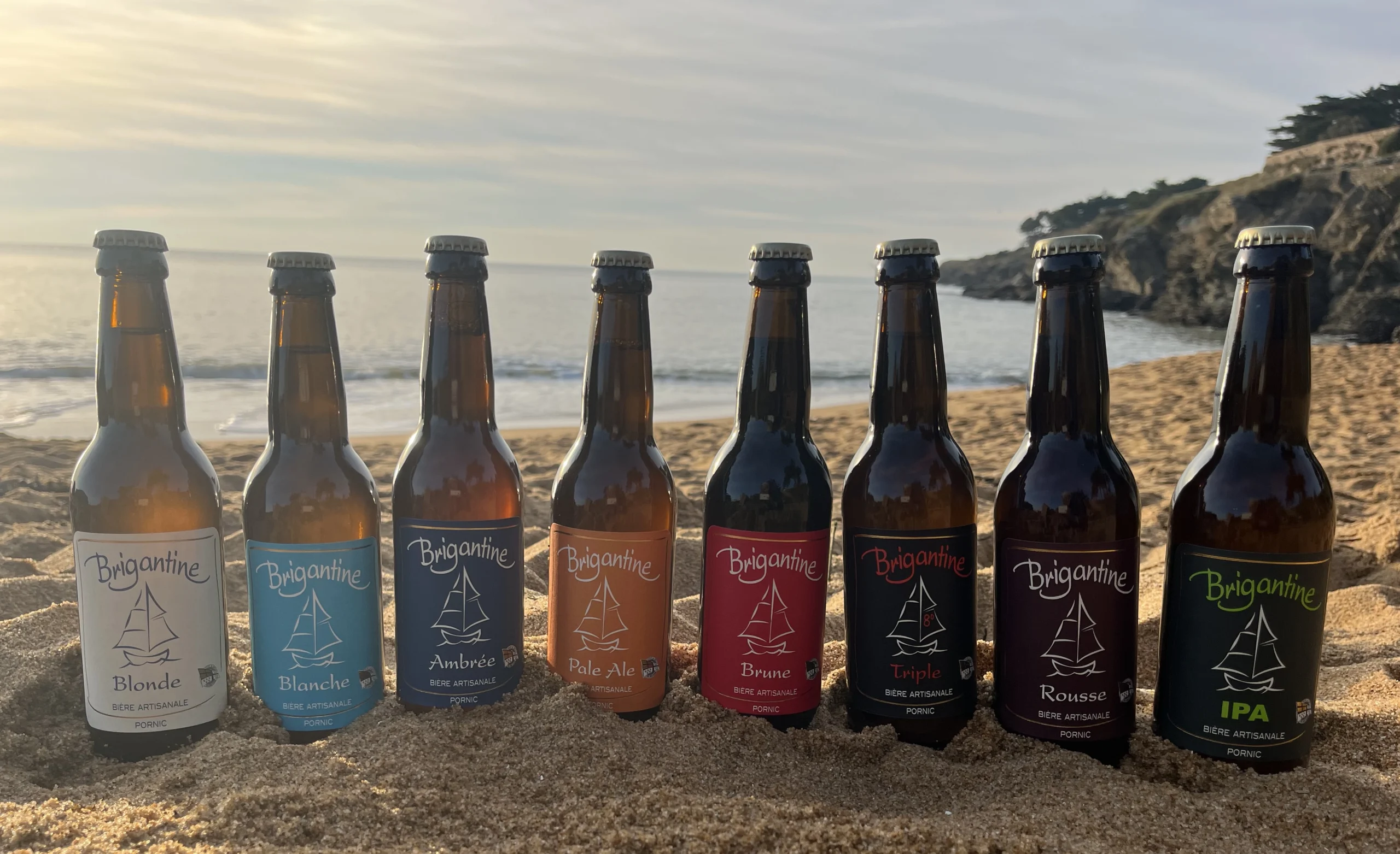 Gamme de huit bières artisanales Brigantine alignées sur une plage avec la mer et des falaises en arrière-plan.