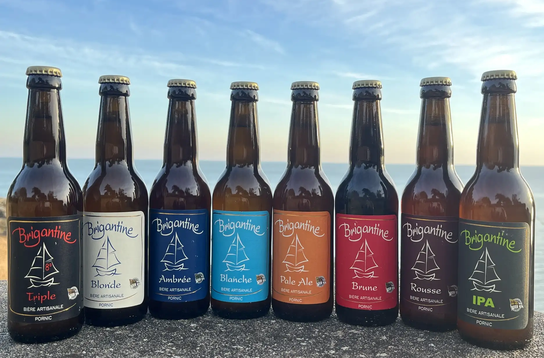 Huit bouteilles de bière artisanale Brigantine alignées face à la mer, présentant différentes variétés allant de la Triple à l'IPA.