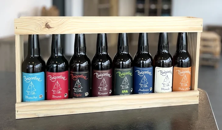 Présentoir en bois clair contenant huit bouteilles de bière artisanale Brigantine alignées, chacune avec une étiquette de couleur différente représentant une variété distincte : Blanche, Brune, Triple, Rousse, IPA, Ambrée, Blonde et Pale Ale.