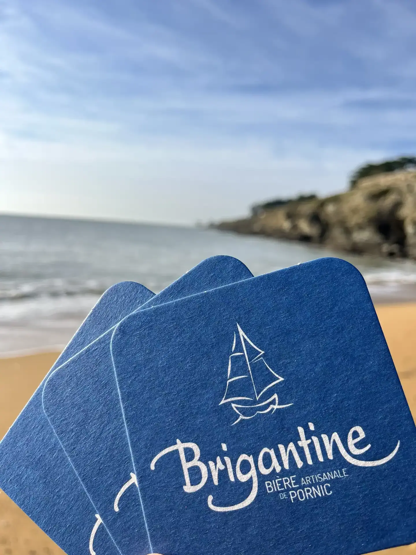 Sous-bocks bleus de la brasserie Brigantine au premier plan avec la plage, la mer et des falaises en arrière-plan.