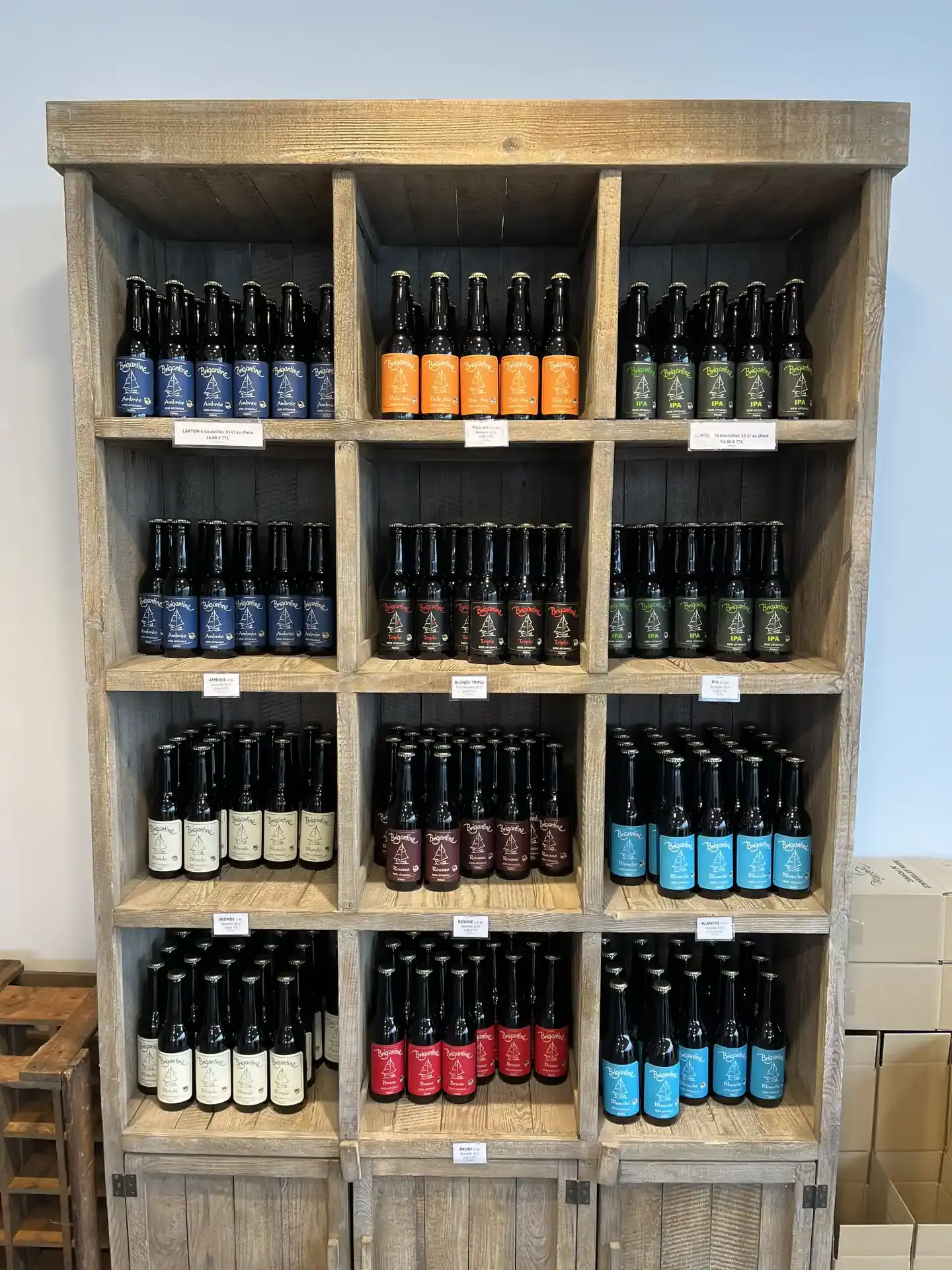 Présentoir en bois à douze compartiments présentant la gamme complète des bières Brigantine avec étiquettes colorées et petites étiquettes de prix.