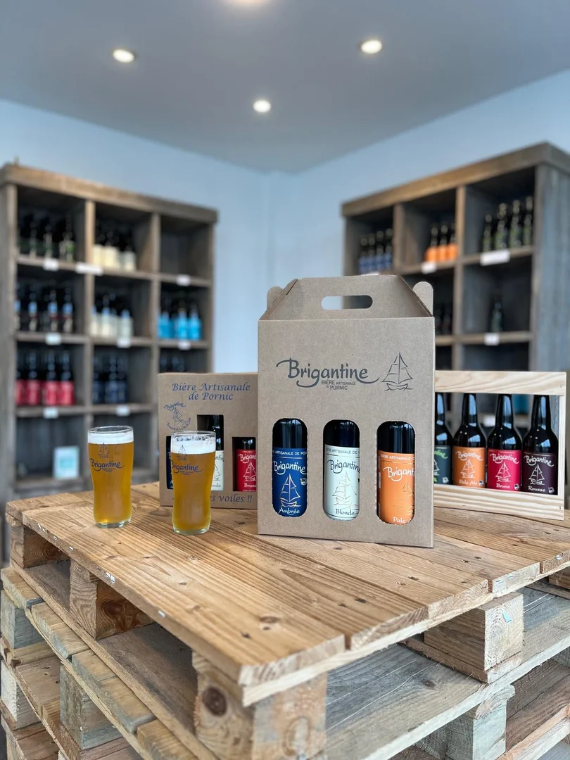 Présentation de bières artisanales Brigantine de Pornic avec un coffret cadeau de trois bouteilles et deux verres remplis de bière blonde sur une table en palettes de bois. En arrière-plan, des étagères en bois contiennent de nombreuses bouteilles de bière de différentes variétés.