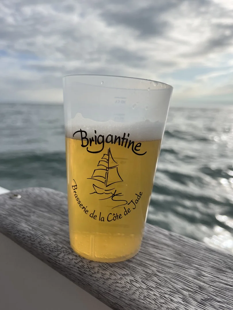 Un verre de bière blonde Brigantine à moitié plein posé sur un rebord en bois avec l'océan et un ciel nuageux en arrière-plan. Le verre porte le logo Brigantine avec un voilier et la mention "Brasserie de la Côte de Jade".