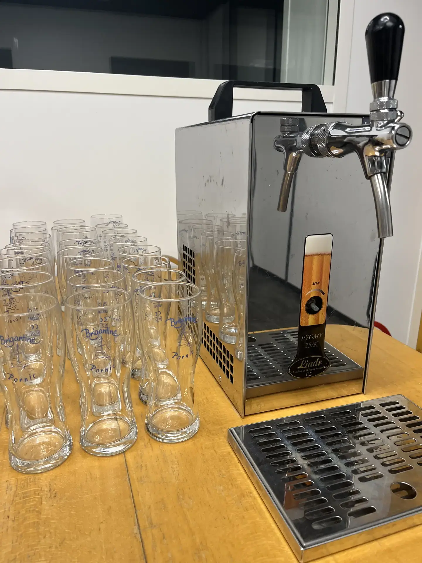 Tireuse à bière professionnelle compacte Lindr Pygmy 25K avec robinet chromé et poignée noire, posée sur un comptoir en bois avec un égouttoir métallique à côté et de nombreux verres vides Brigantine alignés devant.