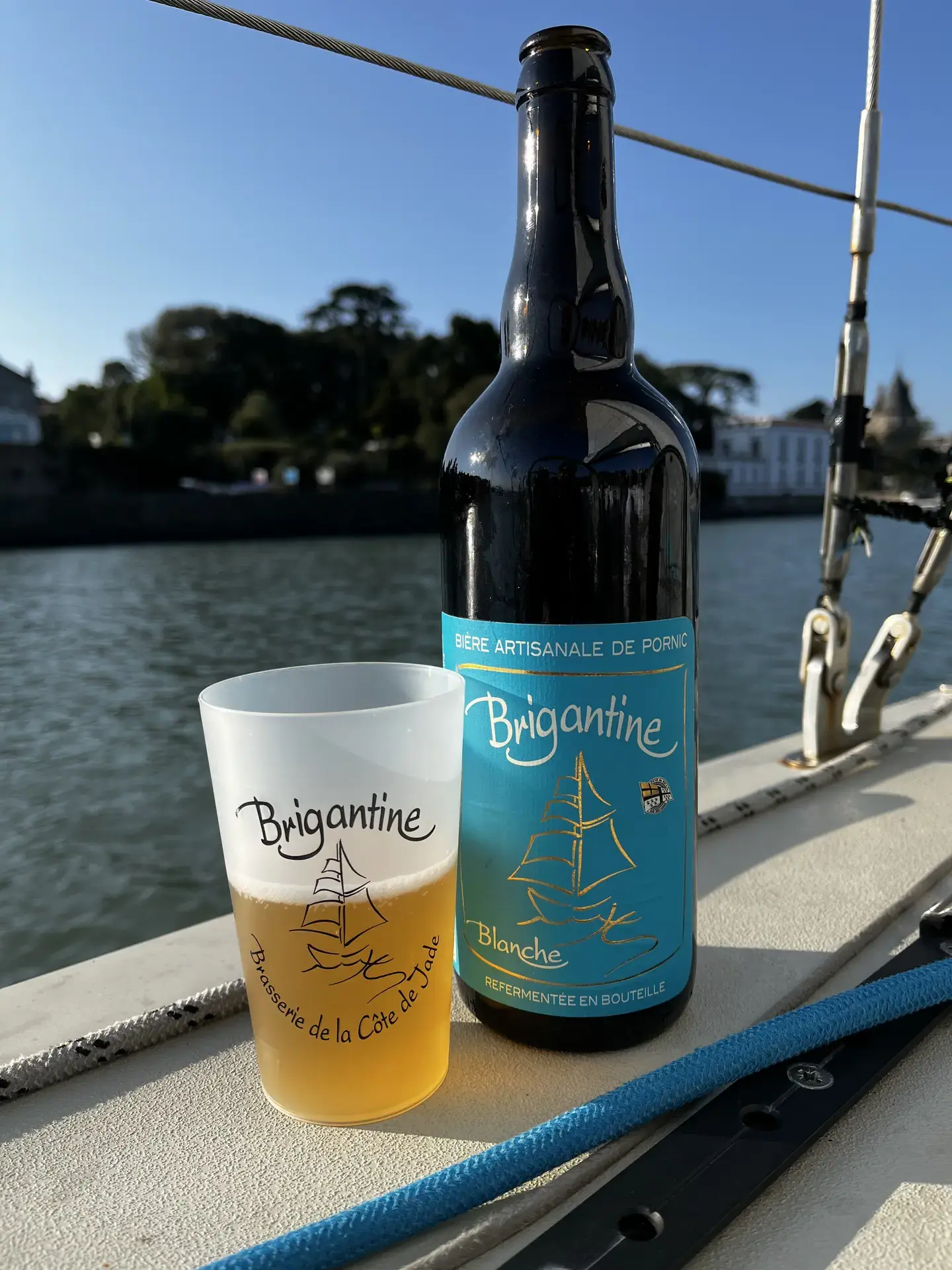 Bouteille de bière Brigantine Blanche et verre à moitié rempli posés sur le pont d'un voilier avec vue sur un port.